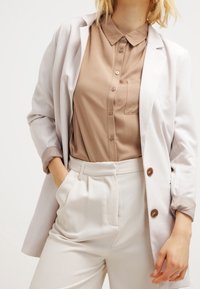 Veste claire portée sur une chemise marron à boutons, associée à un pantalon clair. Comprend un col, une poche et des détails à boutons.