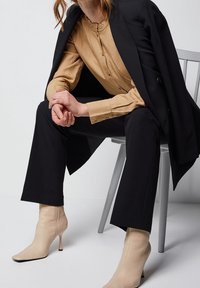 Blusa texturizada beige con cuello con volantes, debajo de un blazer negro ajustado; combinada con pantalones negros y botines beige de punta.