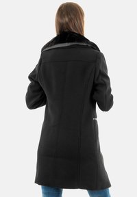 Manteau noir en mélange de laine avec un col en fausse fourrure, coupe ajustée, coutures visibles et longueur atteignant le mi-cuisse, porté sur un jean bleu.