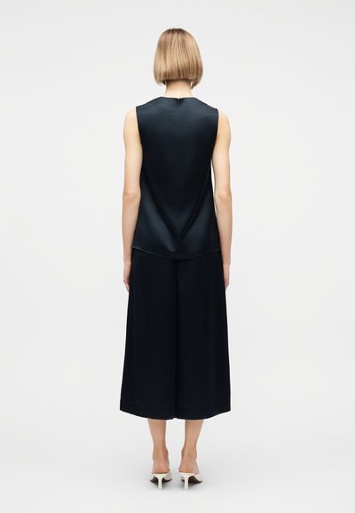 Γυναίκα με κοντά ξανθά μαλλιά που φορεί μαύρο αμάνικο τοπ, μαύρο παντελόνι με φαρδιά μπατζάκια και λευκά παπούτσια με τακούνι, στέκεται με την πλάτη στραμμένη προς τον θεατή.