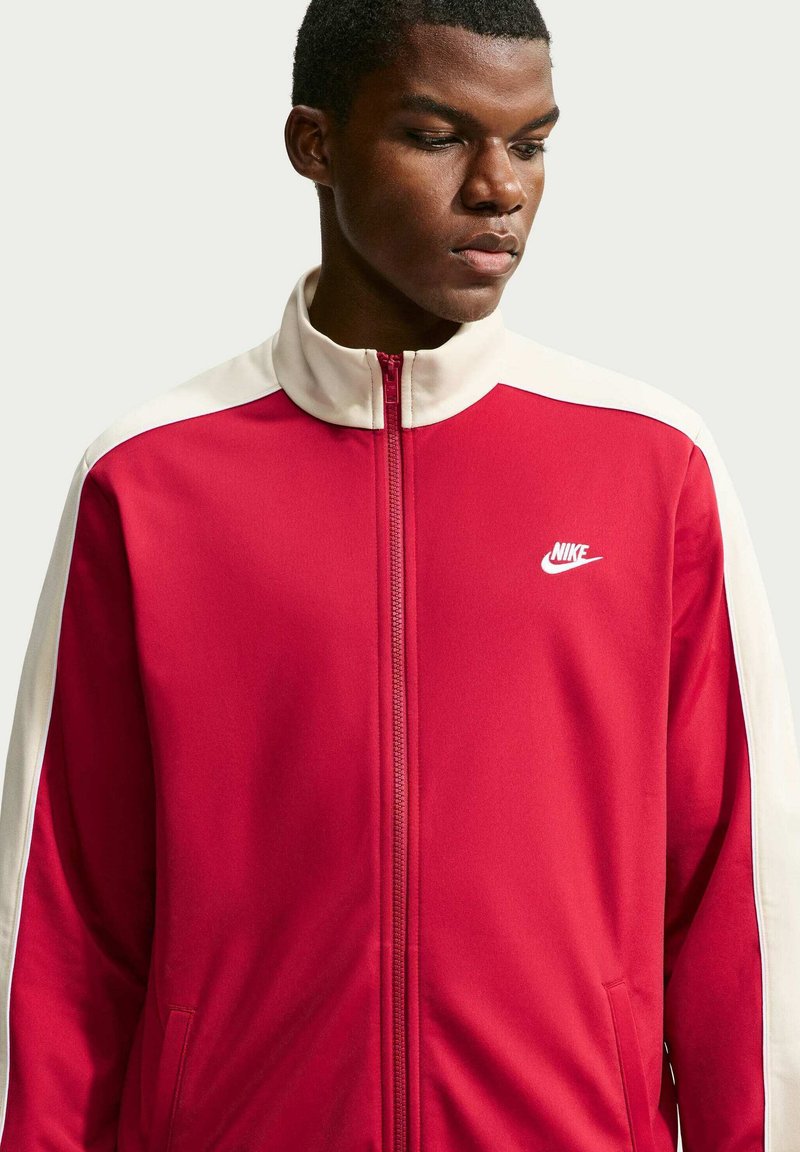 Uomo che indossa una giacca da ginnastica Nike rossa e bianca, guarda in basso e di lato su uno sfondo chiaro uniforme.