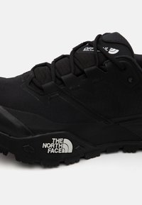 Zapatilla deportiva negra con parte superior de malla texturizada, suela de goma resistente y detalles reflectantes. Presenta un sistema de cordones seguro y un logo.