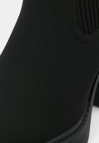 Crne gležnjače od glatkog materijala, s elastičnim bočnim panelima i zaobljenim vrhom. Potplat ima teksturiranu crnu površinu.