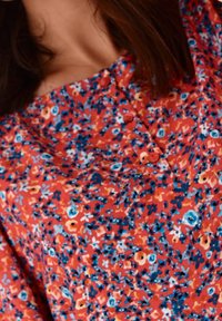 Rød blomstret bluse med et tæt mønster af blå, hvide og gule blomster, med en knapdetalje og et v-halsdesign. Letvægtsstof.