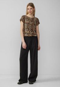Shortsleeve top met luipaardprint in bruin en zwart, gecombineerd met zwarte high-waisted wijde broek. Model draagt platte zwarte schoenen.