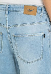 Vaqueros de mezclilla azul claro con una etiqueta de cuero marrón en la parte trasera, que presentan dos bolsillos traseros y detalles de costuras en contraste.