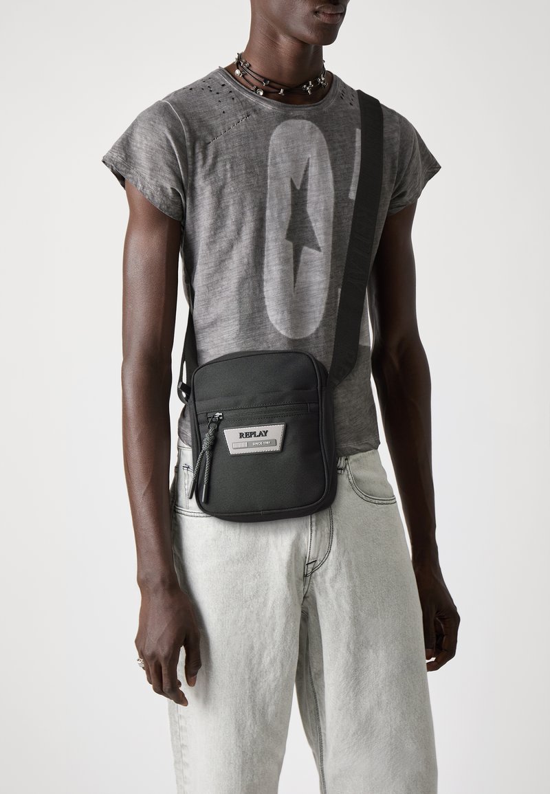 Black crossbody bag made of nylon, featuring a rectangular shape, front zipper pocket, and a silver logo patch. Worn with gray T-shirt and light jeans.