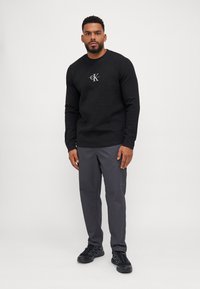 Zwarte crewneck sweatshirt van textuurstof met wit logo op de borst, gecombineerd met grijze broek en zwarte sport schoenen.