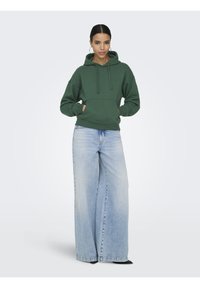 Femme portant un sweat à capuche vert foncé, les mains dans la poche avant, et un jean bleu clair à jambes larges, sur un fond clair uni.