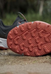 Chaussure de trail noire et grise avec une semelle épaisse rouge à crampons, posée sur un sol de terre à l'extérieur.