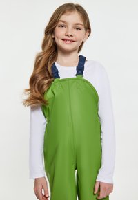 Žalios vandeniui atsparios pertraukos su reguliuojamomis mėlynomis petnešomis, dėvimos ant baltos ilgomis rankovėmis marškinės. Lygus paviršius, sukurtas lauko naudojimui.