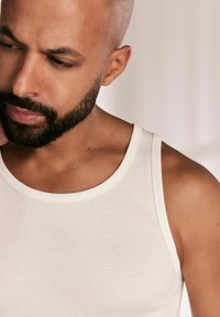 Weißes, geripptes Tanktop mit rundem Halsausschnitt, getragen von einer Person mit kurzem Haar und Bart. Glatte Textur, figurnahe Passform.