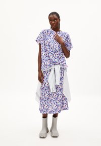 Frau steht in einem lila Blumenkleid mit einem hellgrauen Hemd, das um die Taille gebunden ist, und grauen klobigen Stiefeln vor einem weißen Hintergrund.