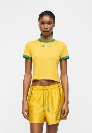 Kobieta w żółtej krótkiej koszulce z zielonym obszyciem i żółtych szortach ze zielonymi sznureczkami, stojąca prosto na jednolitym tle.