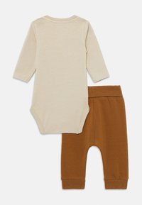 Beige Babykombination mit langen Ärmeln und Druckknöpfen, kombiniert mit braunen, gerippten Babyhosen, flach auf einem weißen Hintergrund ausgelegt.