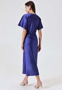 Abito blu in satin con maniche a sbuffo corte, lunghezza media e dettaglio a chiave sulla schiena, abbinato a scarpe con tacco beige e una consistenza liscia.