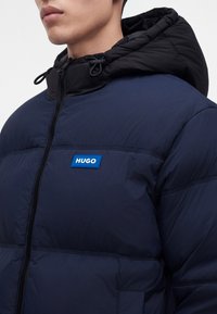 Homme portant une doudoune bleu marine avec une capuche noire et un patch bleu marqué "HUGO" sur la poitrine.