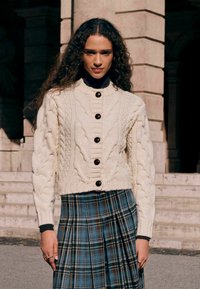Cream cable-knit cardigan with a high collar and brown buttons, paired with a blue plaid pleated skirt. Textured knit and fitted design.