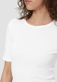 Witte katoenen t-shirt met korte mouwen, ronde hals en een gladde textuur, waarbij een deel van een schouder en nek in het beeld zichtbaar is.