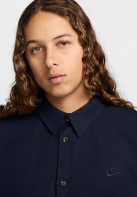Camisa de colarinho azul-marinho com botão à frente, apresentando botões castanhos e um logótipo da Nike em azul escuro bordado no lado esquerdo do peito.