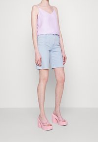 Kobieta w jasnofioletowej bezrękawnikowej bluzce, jasnoniebieskich szortach dżinsowych i różowych sandałach na platformie na wysokim obcasie, stojąca na jednolitym tle.