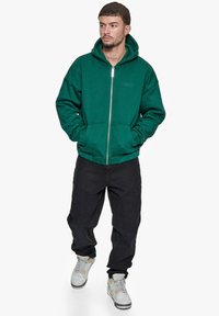 Grøn zip-up hættetrøje med afslappet pasform, prydet med ensfarvet design, kængurulomme og subtil logo-detalje på brystet.