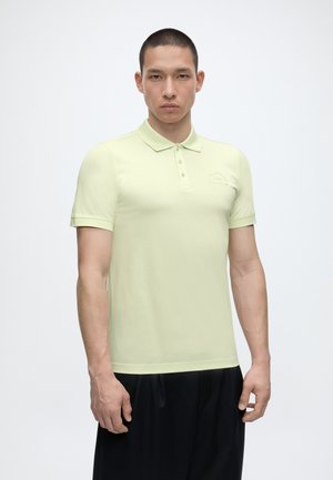 Mladý muž s krátkymi vlasmi v svetložltej polokošeli s krátkym rukávom a čiernych nohaviciach stojaci na jednoduchom pozadí.