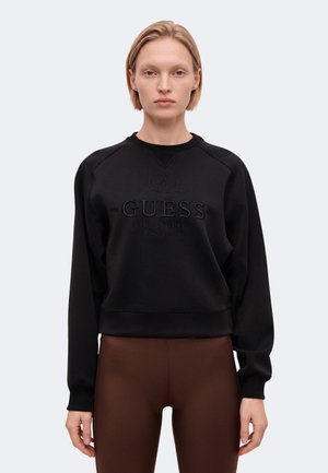 Модел с къса руса коса носи черен суитшърт с логото на Guess и кафяв панталон, застанал на светъл фон.