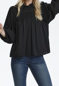 Svart långärmad blus med hög halsringning, smockdetaljer över bysten och en avslappnad passform, ihop med blå denimjeans.