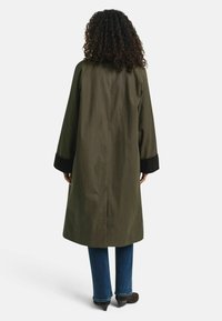 Person mit lockigem Haar, der einen langen olivgrünen Mantel mit schwarzen Ärmeln, blaue Jeans und schwarze Absatzschuhe trägt, von hinten gesehen.