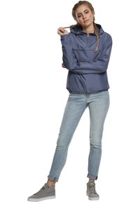 Giubbotto a vento blu chiaro con cappuccio, colletto con zip e tasca frontale, abbinato a jeans in denim chiaro e sneakers grigie.