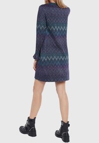 Vestito viola a zigzag con maniche lunghe, caratterizzato da un collo rotondo e una silhouette dritta. Indossato con stivaletti neri con borchie.