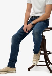 Hombre con zapatillas beige, vaqueros azules, camiseta blanca y reloj sentado en un taburete de madera oscura frente a un fondo blanco.