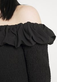 Top nero scollato con spalle scoperte in tessuto testurizzato, con scollatura arricciata a ruffles e dettagli smock lungo il corpo.