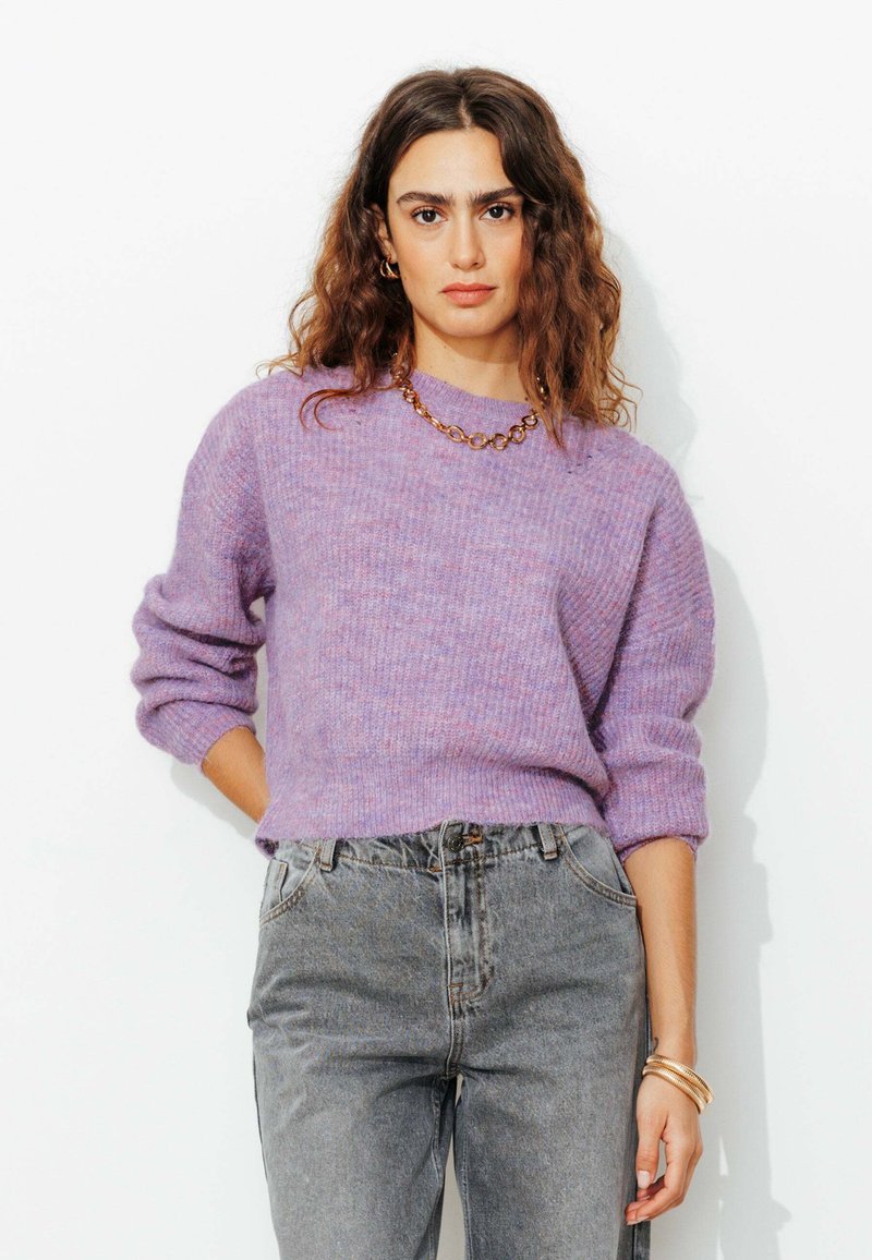 Femme aux cheveux bruns ondulés portant un pull en tricot lavande, un jean gris taille haute, un collier en chaîne dorée et des bracelets, se tenant contre un mur blanc.