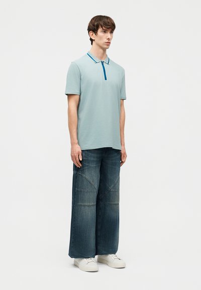 Jeune homme debout portant un polo à manches courtes bleu clair, un jean large foncé avec des détails cousus, et des baskets blanches sur un fond uni.