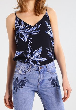 Nainen, joka pukeutunut mustaan hihattomaan toppiin, jossa on sininen kukkakuvio, ja vaaleansiniset korkeavyötäröiset farkut, joissa on tummansininen kukkakirjailu reisissä.