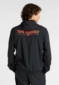 Schwarze Kapuzenjacke mit glatter Textur, ausgestattet mit einem auffälligen roten "New Balance"-Logo auf dem Rücken. Elastischer Saum und Bündchen für eine bequeme Passform.