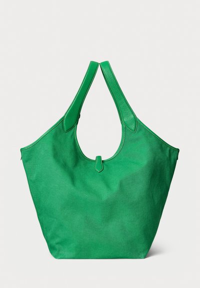 Liela zaļa auduma soma ar platām ādas rokturiem un auduma korpusu, ar mazu siksniņu detaļu centrālajā atvērumā uz vienkrāsaina fona.