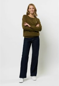 Olivnozeleni pulover s krožnim izrezom in rebrastimi manšetami, v kombinaciji s temno modrimi širokimi hlačami in belimi supergami. Model z prekriženimi rokami.