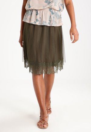 Olivgrüner Tüllrock mit Spitzenbesatz am Saum. Das mehrlagige Design sorgt für Struktur. Kombiniert mit einer Bluse mit Blumenmuster und Sandalen.