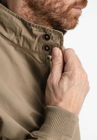 Chaqueta beige con un cuello alto, que presenta dos botones oscuros y puños acanalados. El material parece suave, con un diseño ligero.