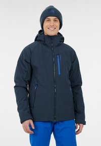 Marineblauwe gewatteerde jas met ritssluitingen, capuchon en accenten in felblauw, gecombineerd met blauwe broek. De textuur lijkt glad en duurzaam.