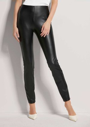Vrouw draagt strakke zwarte leggings van imitatieleren stof, gecombineerd met witte pumps met een spits toelopende neus en een wit topje, staand tegen een lichte achtergrond.