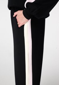 Pantalon de jogging noir avec une bande latérale en rose pâle et blanc. Matière douce, avec une poche inclinée sur la hanche et des poignets côtelés.