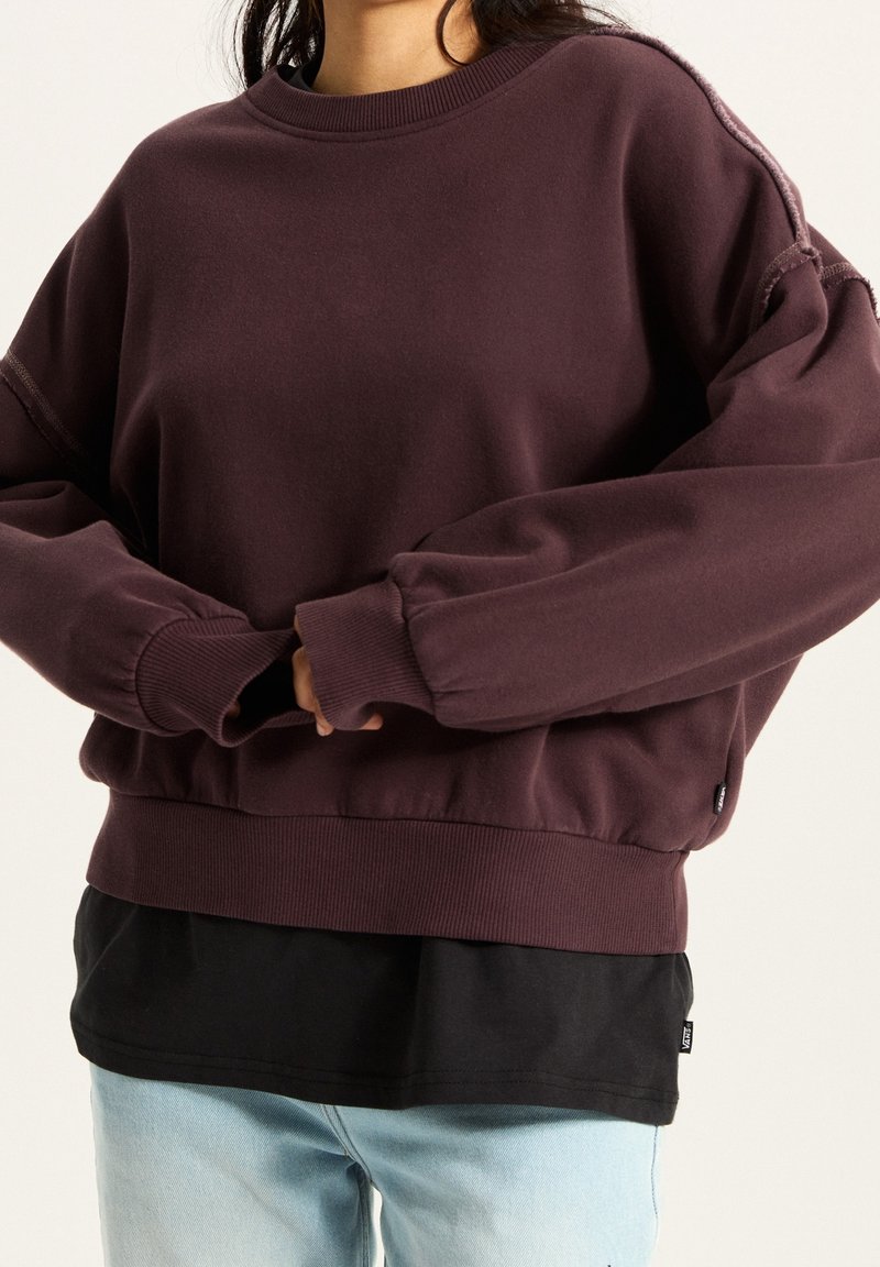 Personne portant un sweat oversize violet foncé sur une chemise noire avec un jean bleu clair, mains jointes à la taille.