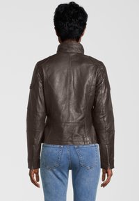 Braune Leder-Motorradjacke mit hohem Kragen, gesteppten Details an den Ärmeln und Reißverschlussmanschetten. Getragen über hellblauen Jeans.