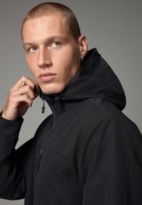 Giacca nera con cappuccio realizzata in un materiale liscio e traspirante. Presenta una chiusura con zip frontale, un cappuccio regolabile con cordino e una tasca sul petto con zip.