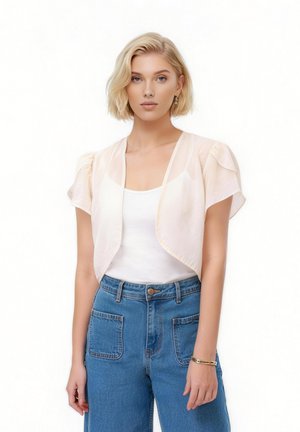 Ung kvinde med kort blondt hår iført en gennemsigtig cremefarvet kortærmet shrug over en hvid tanktop og højtaljede blå jeans.