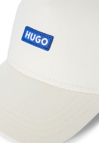 Witte pet met een gestructureerd ontwerp, een blauw rechthoekig vak met "HUGO" in witte letters en zichtbare stikdetails.